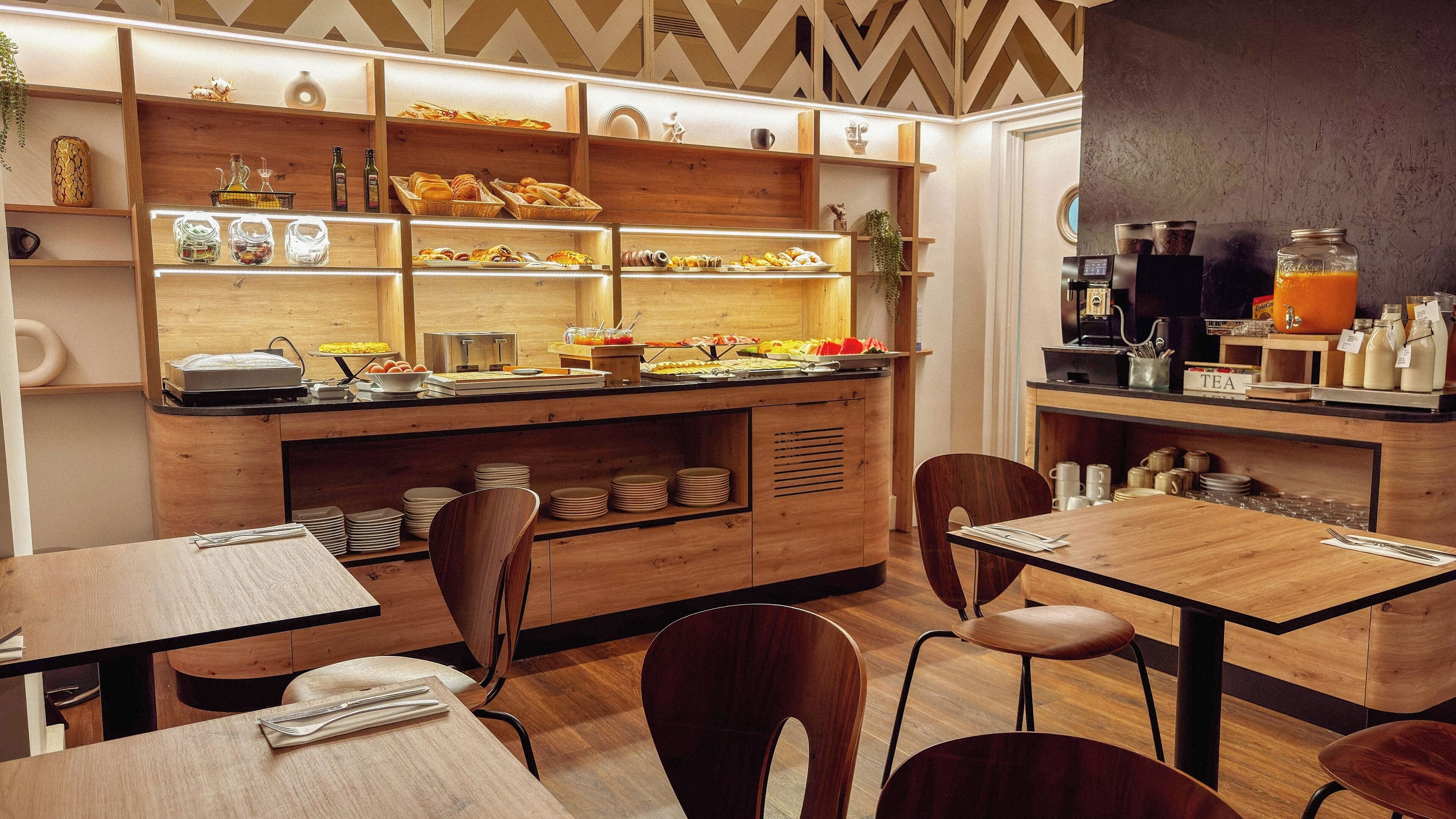 Un café moderno con mobiliario de madera, una estación de café y diversas opciones de comida exhibidas en estanterías.