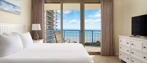 A hotel room with a large bed, a TV, a balcony with a view of the ocean, and a ceiling fan.