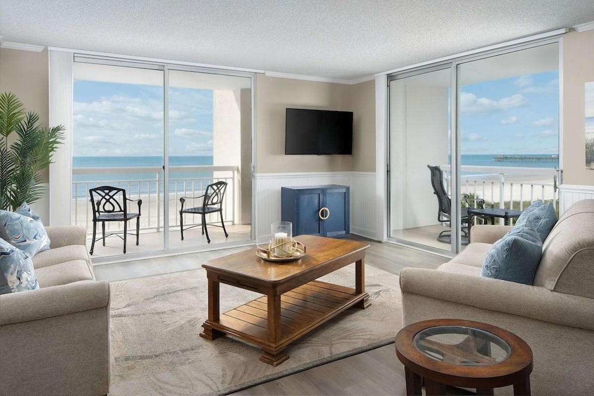 A living room with a sofa, a coffee table, and a TV. There is a balcony with a view of the beach.