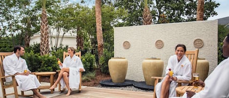 A group of people in white robes seated on wooden chairs in an outdoor setting with a wooden deck, surrounded by greenery and a decorative wall with circular patterns.