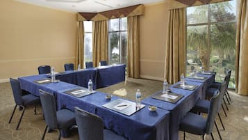 A conference room with a long table, chairs, water bottles, and a view of palm trees through large windows.
