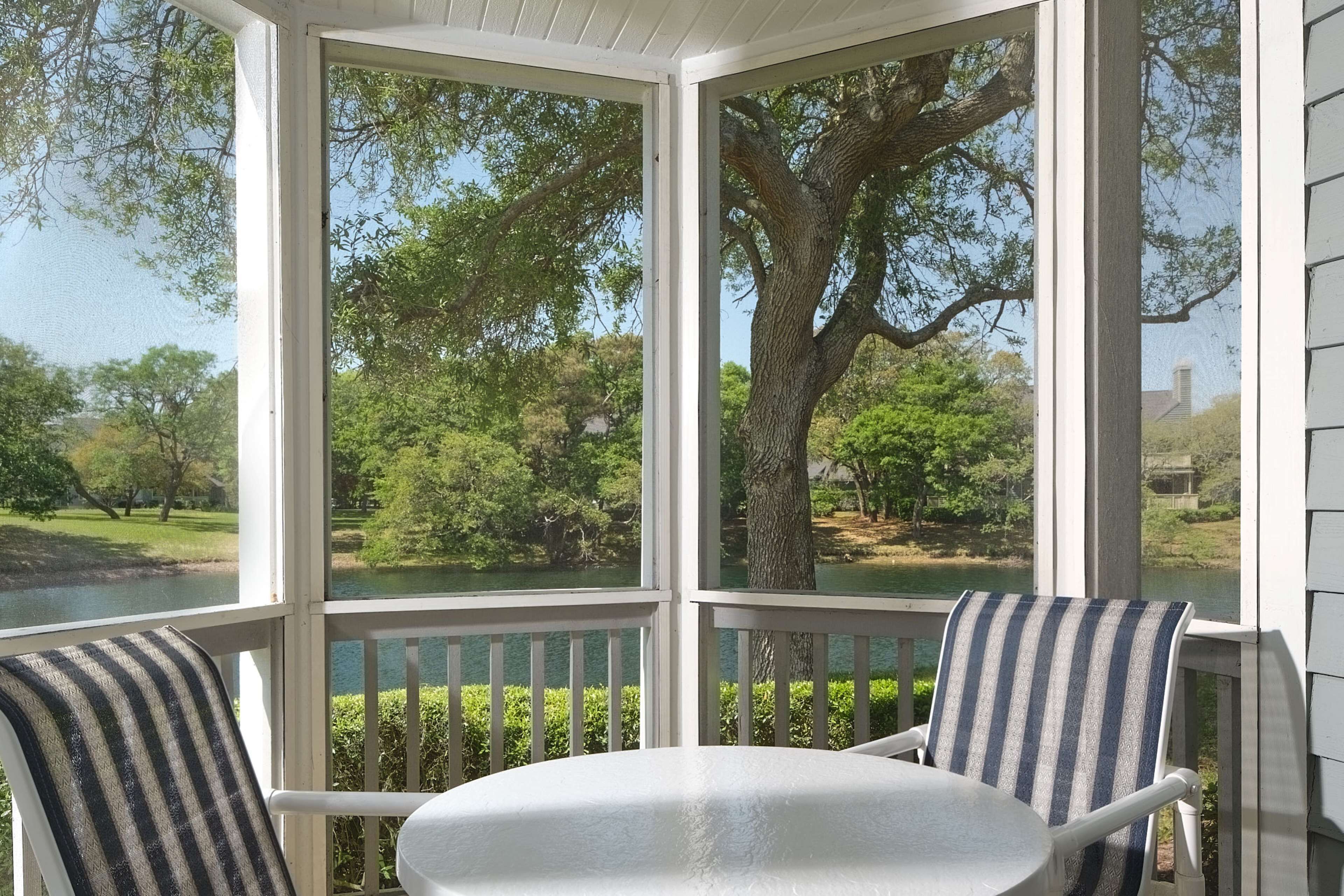 A covered outdoor seating area with a table and chairs, overlooking a natural landscape with trees and a body of water.