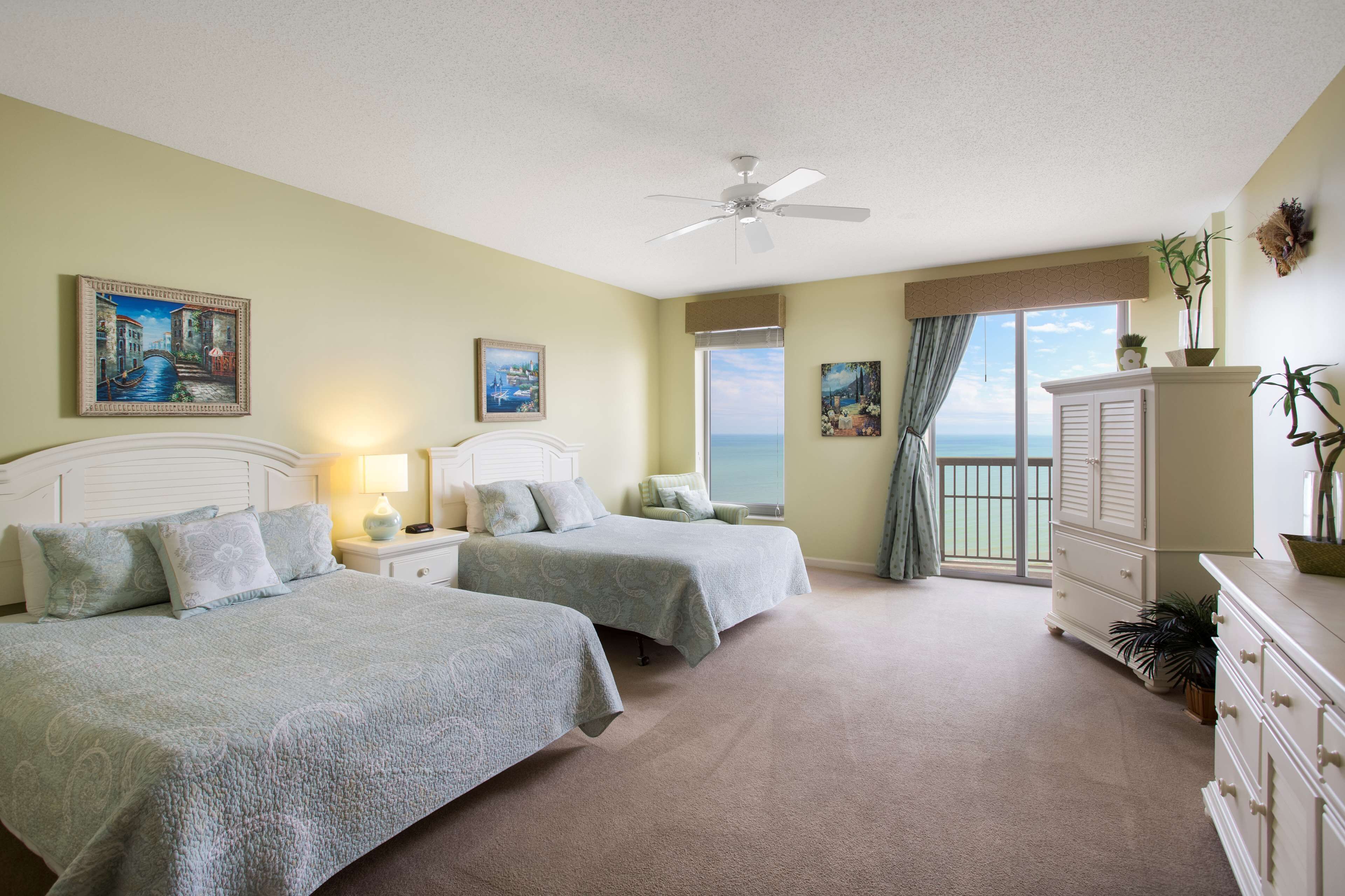 A bedroom with two beds, a ceiling fan, a nightstand with a lamp, a dresser, and a view of the ocean through a sliding door.