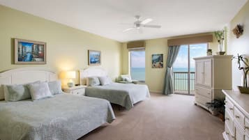 A bedroom with two beds, a ceiling fan, a nightstand with a lamp, a dresser, and a view of the ocean through a sliding door.