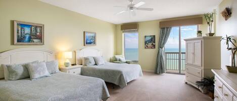 A bedroom with two beds, a ceiling fan, a nightstand with a lamp, a dresser, and a view of the ocean through a sliding door.