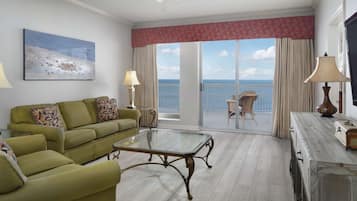 A hotel room with a green sofa, a glass coffee table, a dining table, a TV, and a balcony with a view of the sea.