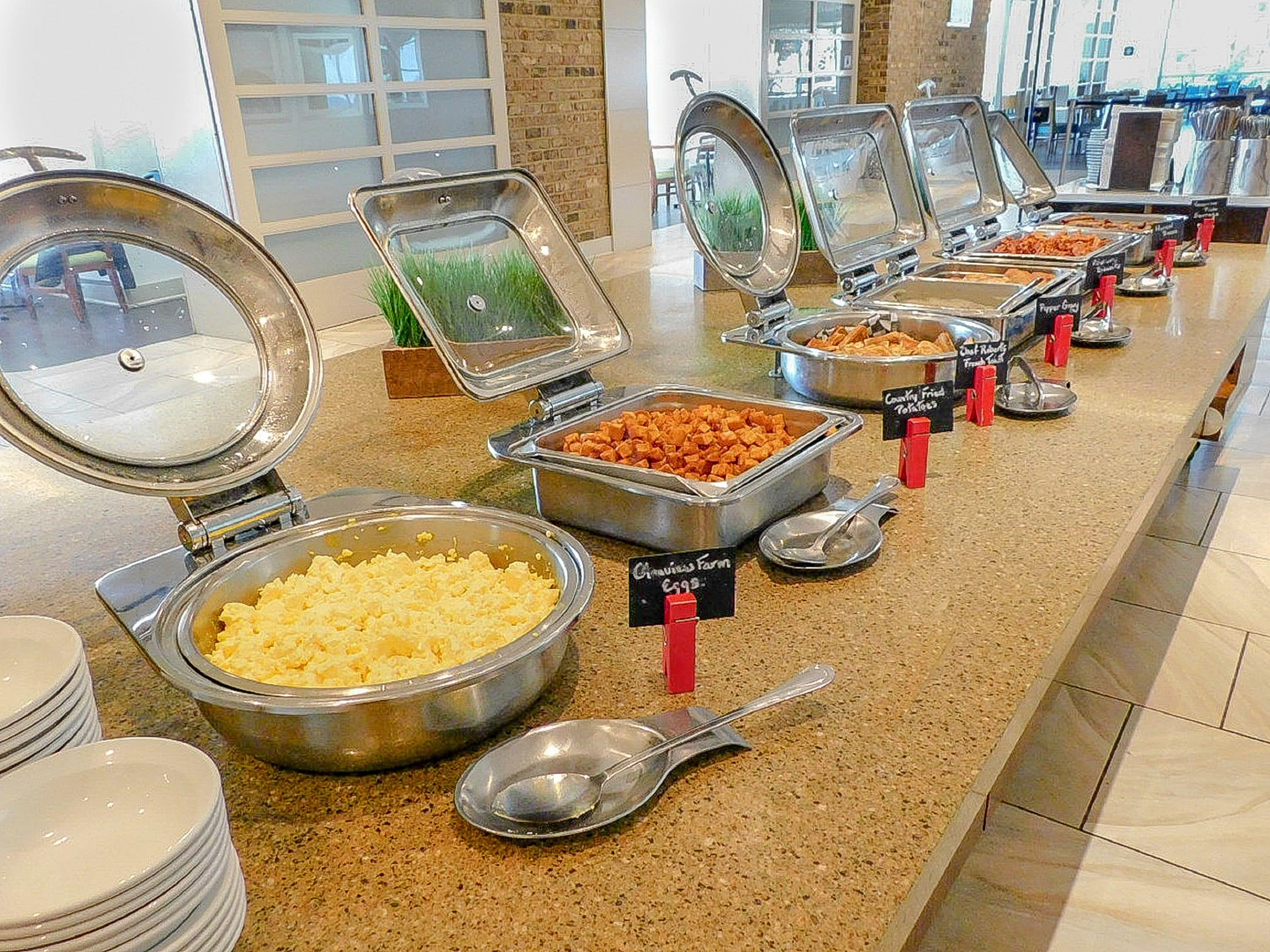 Un buffet proposant une variété de plats, notamment des œufs brouillés, des pommes de terre sautées à la poêle, et d’autres mets, avec des ustensiles de service et des étiquettes indiquant les prix.