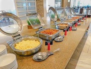 Un buffet proposant une variété de plats, notamment des œufs brouillés, des pommes de terre sautées à la poêle, et d’autres mets, avec des ustensiles de service et des étiquettes indiquant les prix.