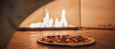 Pizza in einem Holzofen, im Hintergrund sind Flammen zu sehen.