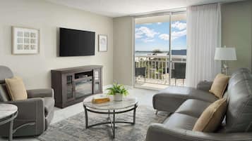 A modern living room with a sofa, armchair, and a small table with a plant. A flat-screen TV is mounted on the wall, and there is a balcony with a view of the sky and clouds.