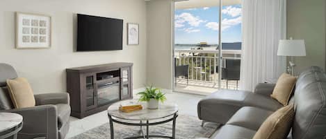 Una sala de estar moderna con un sofá, un sillón y una mesita con una planta. Un televisor de pantalla plana está montado en la pared, y hay un balcón con vistas al cielo y nubes.