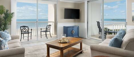 A living room with a large sliding glass door leading to a balcony with a view of the ocean. There is a wooden coffee table, a sofa, and a TV mounted on the wall.