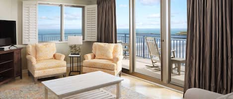 A spacious living room with two patterned armchairs, a white coffee table, and a sofa. A television is on a wooden stand, and a small table with a lamp is in the corner. The room has large windows with curtains, offering a view of the sea and a bridge.