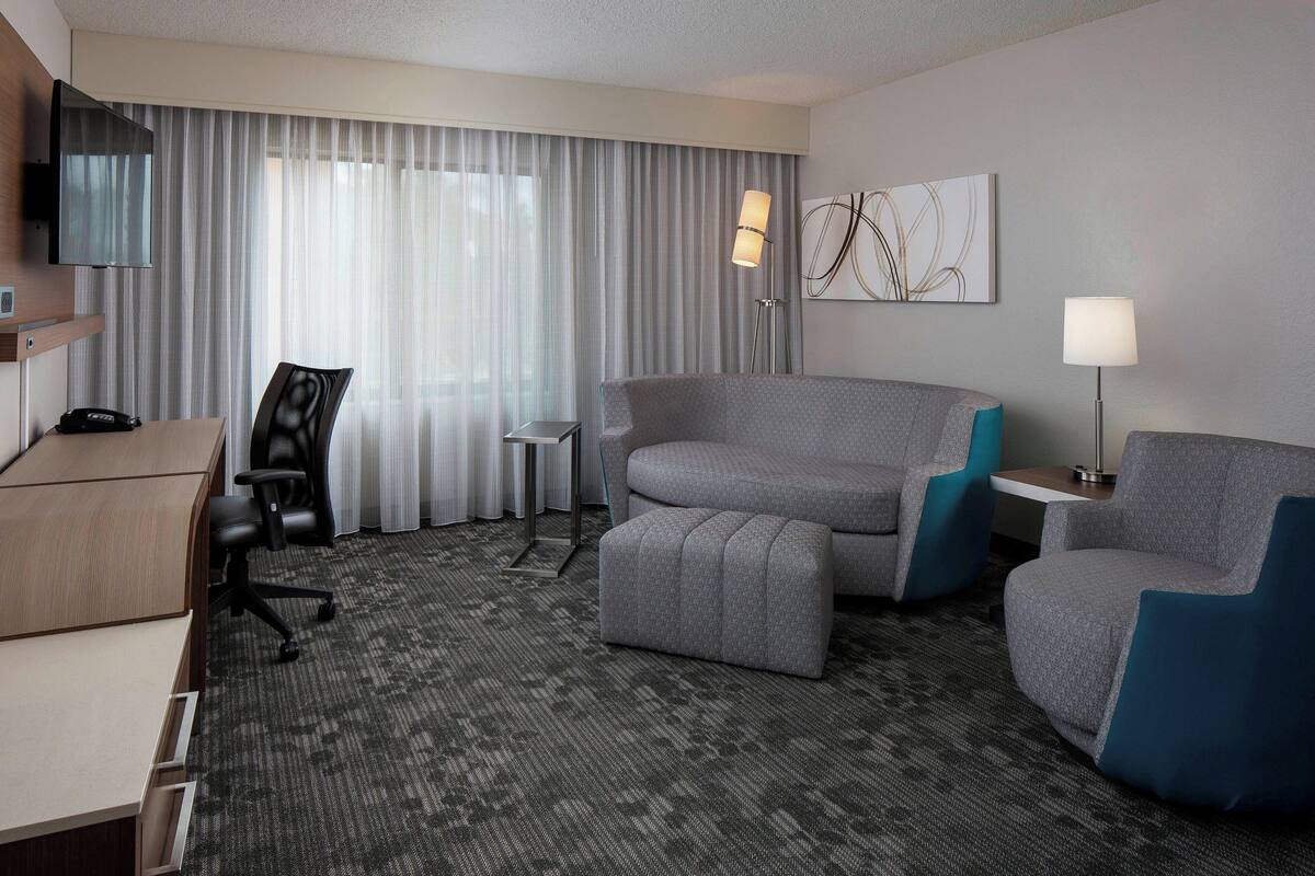 A hotel room with a desk, two armchairs, a sofa, a small table, a lamp, and a TV mounted on the wall.