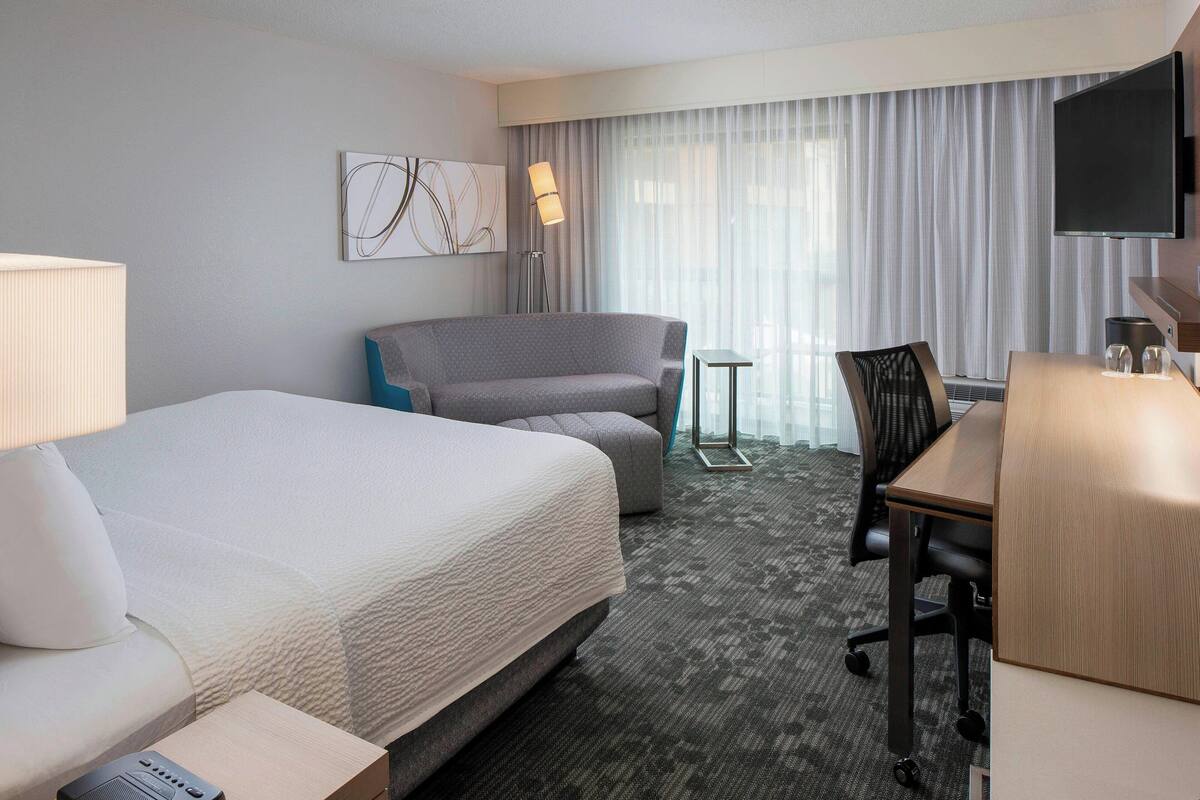 A hotel room with a large bed, a desk, a chair, a TV, and a window with curtains.