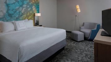 A hotel room with a large bed, a desk with a flat-screen TV, a chair, and a floor lamp.