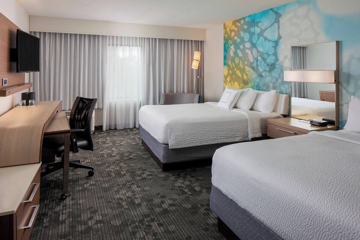 A hotel room with two beds, a desk, a chair, a TV, and a large window with curtains.