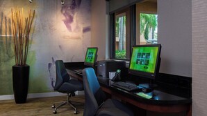 A modern office with two workstations, each with a computer monitor, keyboard, and mouse. There is a tall vase with branches on the left, and a window providing a view of palm trees outside. - Courtyard by Marriott Daytona Beach Speedway/Airport (Daytona Beach)