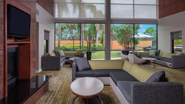 A modern lounge area with a large sectional sofa, a round coffee table, and a flat-screen TV mounted on the wall. - Courtyard by Marriott Daytona Beach Speedway/Airport (Daytona Beach)