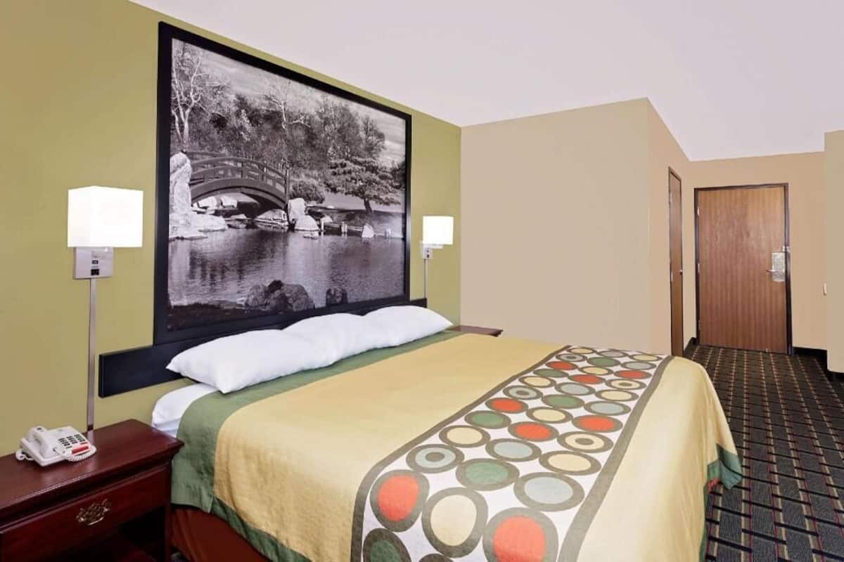 Habitación de hotel con cama, mesita de noche con teléfono, lámpara de pared y una puerta, con una colcha estampada.