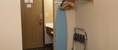 A hotel room with a wooden door, a blue ironing board, a hanging rack with clothes, and a crutch.