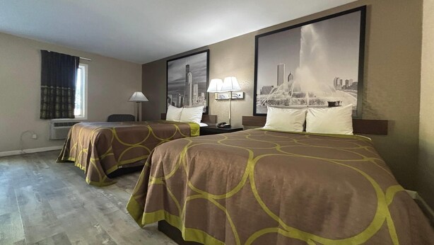 A hotel room with two beds, a large window with curtains, a wall-mounted lamp, and two framed cityscape pictures.