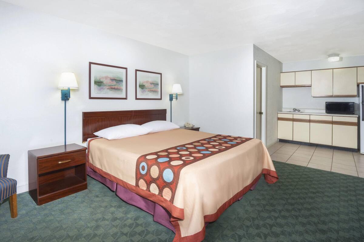 A hotel room with a bed, bedside table, lamp, and a kitchenette area.
