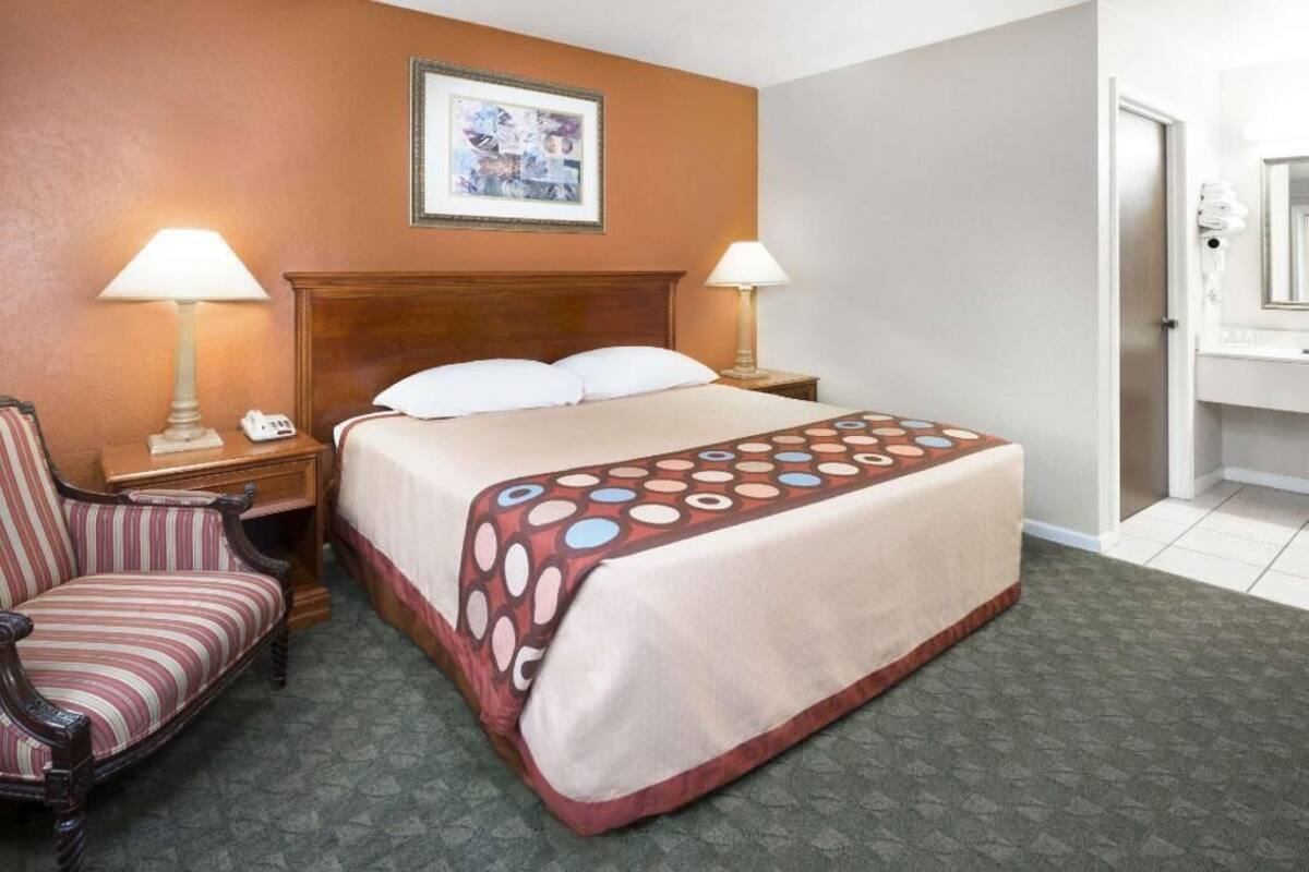 A hotel room with a bed, two bedside lamps, a chair, a framed picture on the wall, and a bathroom visible through an open door.