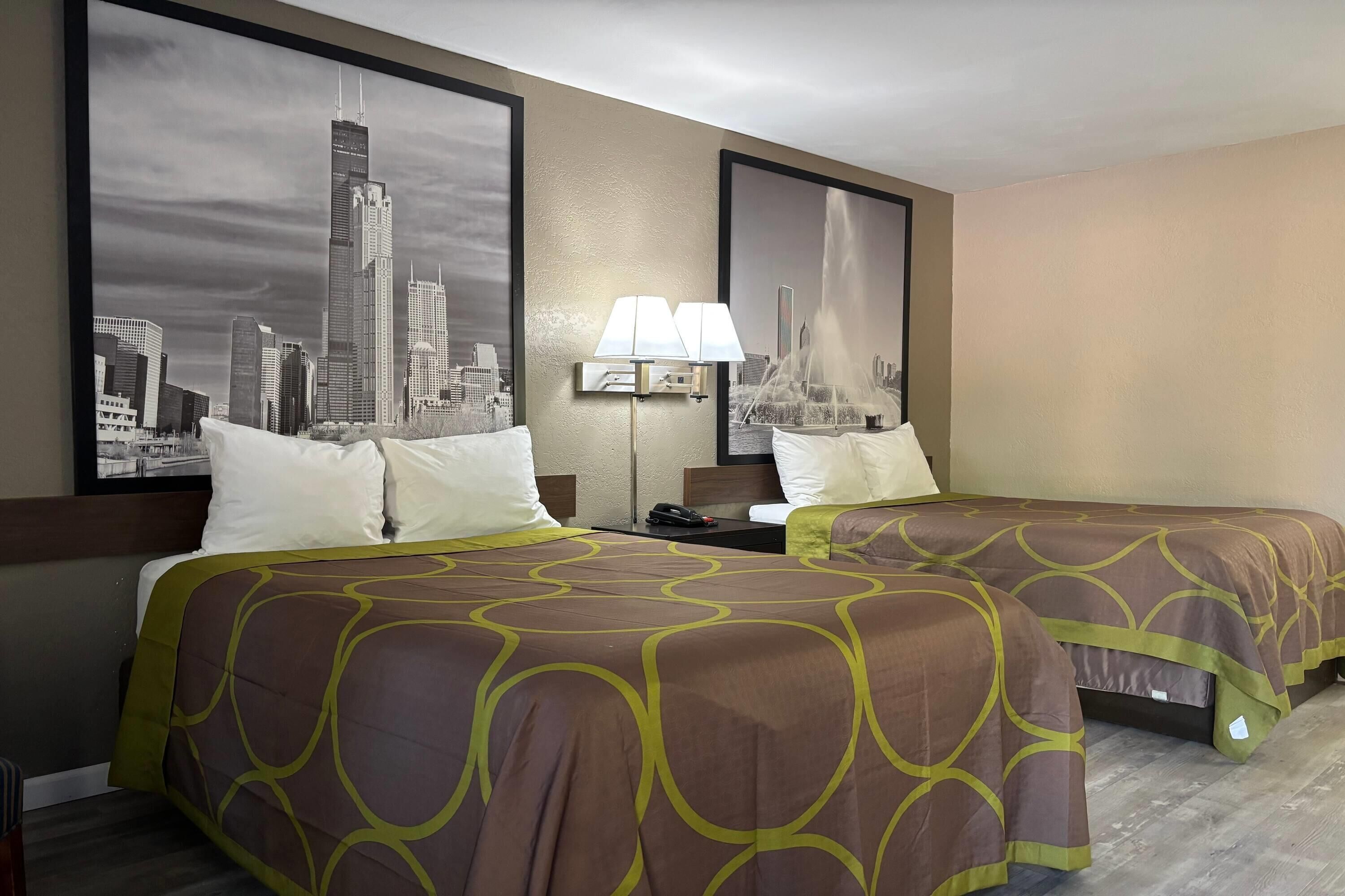 Quarto de hotel com duas camas, uma mesinha de cabeceira, uma luminária e duas grandes fotografias emolduradas de paisagem urbana na parede.