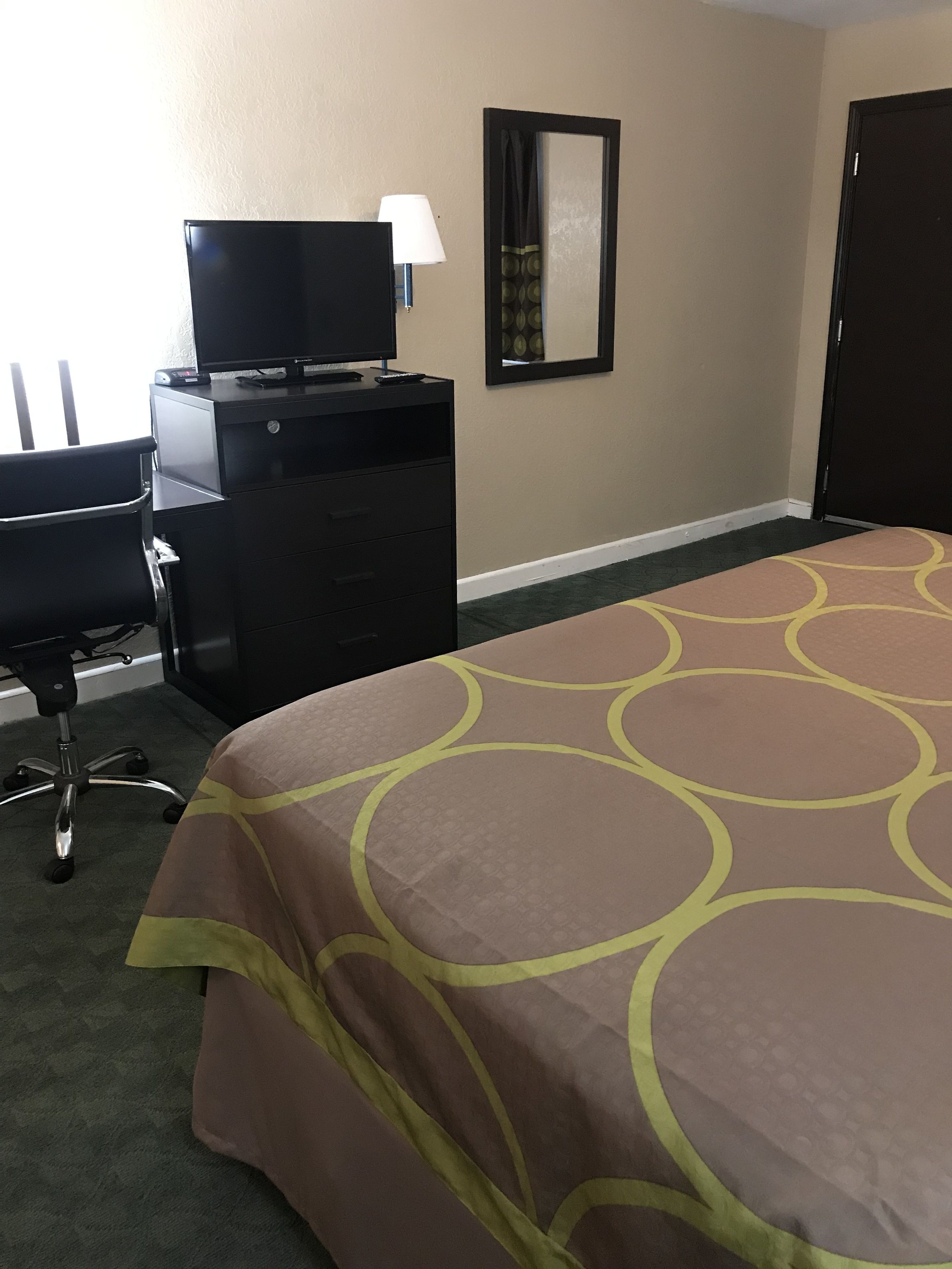 A hotel room with a bed, a desk with a computer, a chair, a lamp, and a door.