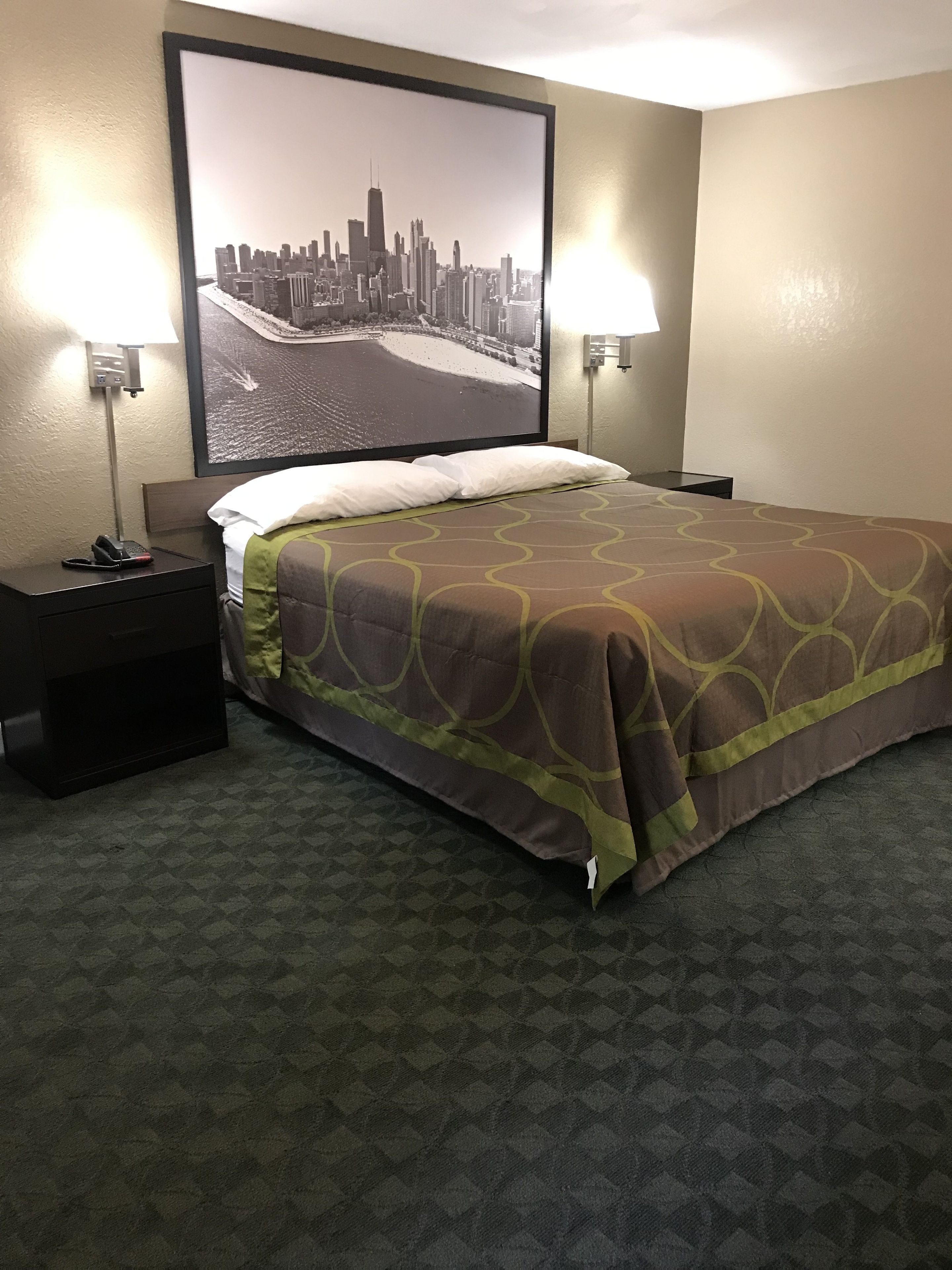Habitación de hotel con una cama, dos mesitas de noche, una mesita auxiliar, un cartel grande con vistas a la ciudad en la pared y una alfombra estampada.