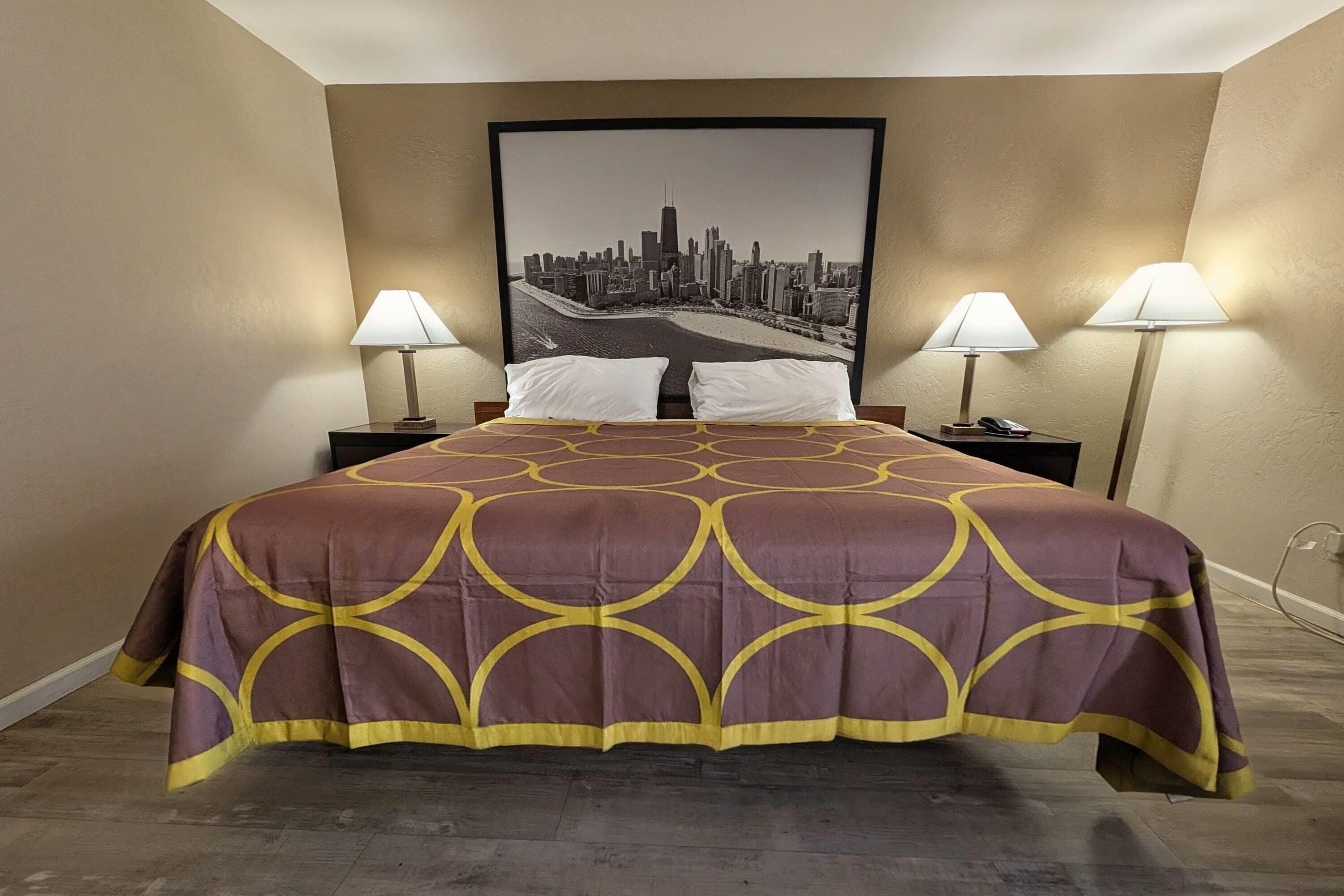 A hotel room with a large bed, two bedside tables with lamps, a cityscape painting on the wall, and a patterned bedspread.