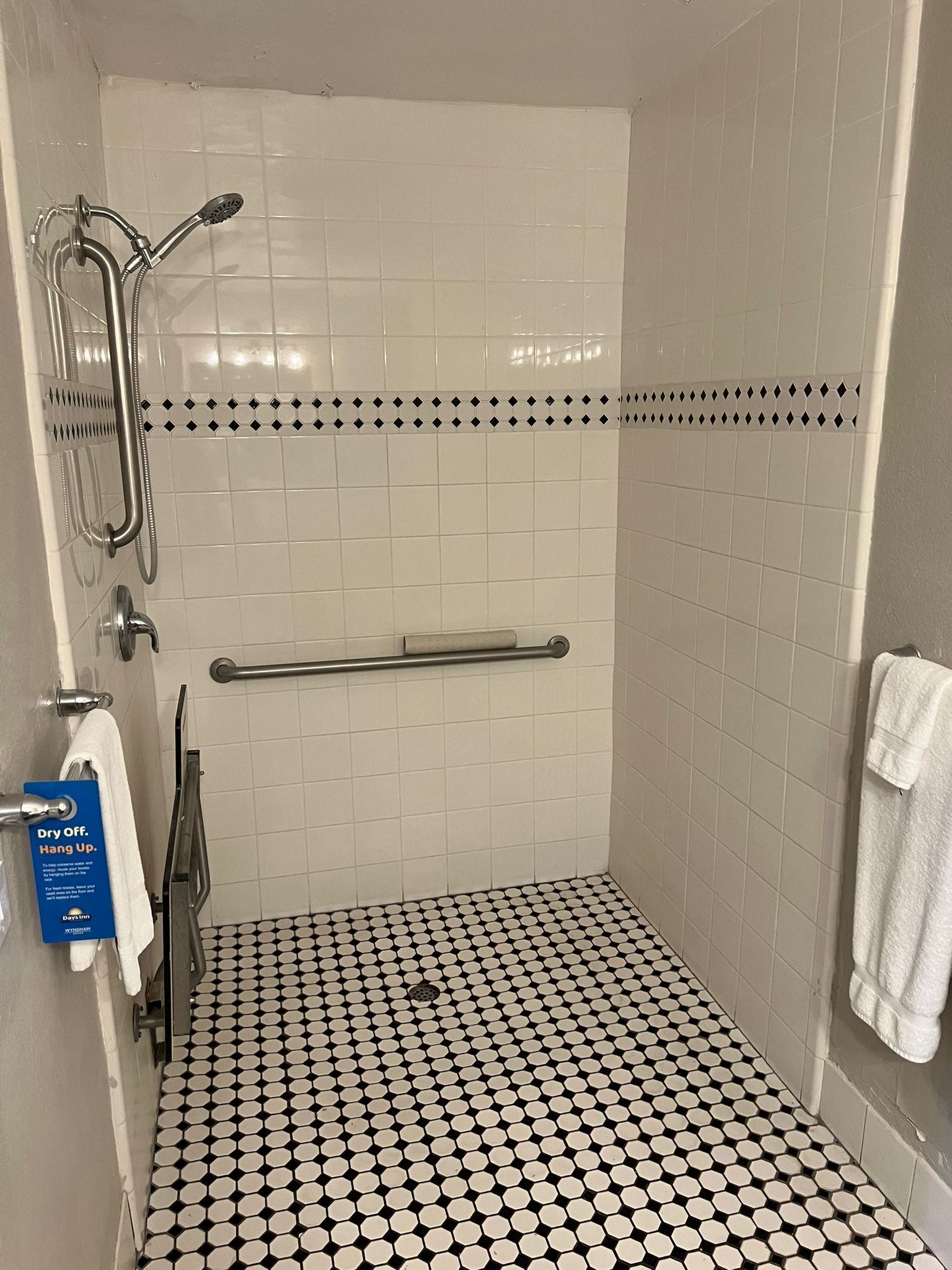 Um chuveiro pequeno, revestido com azulejos, com piso xadrez preto e branco, uma barra de apoio e um porta-toalhas.