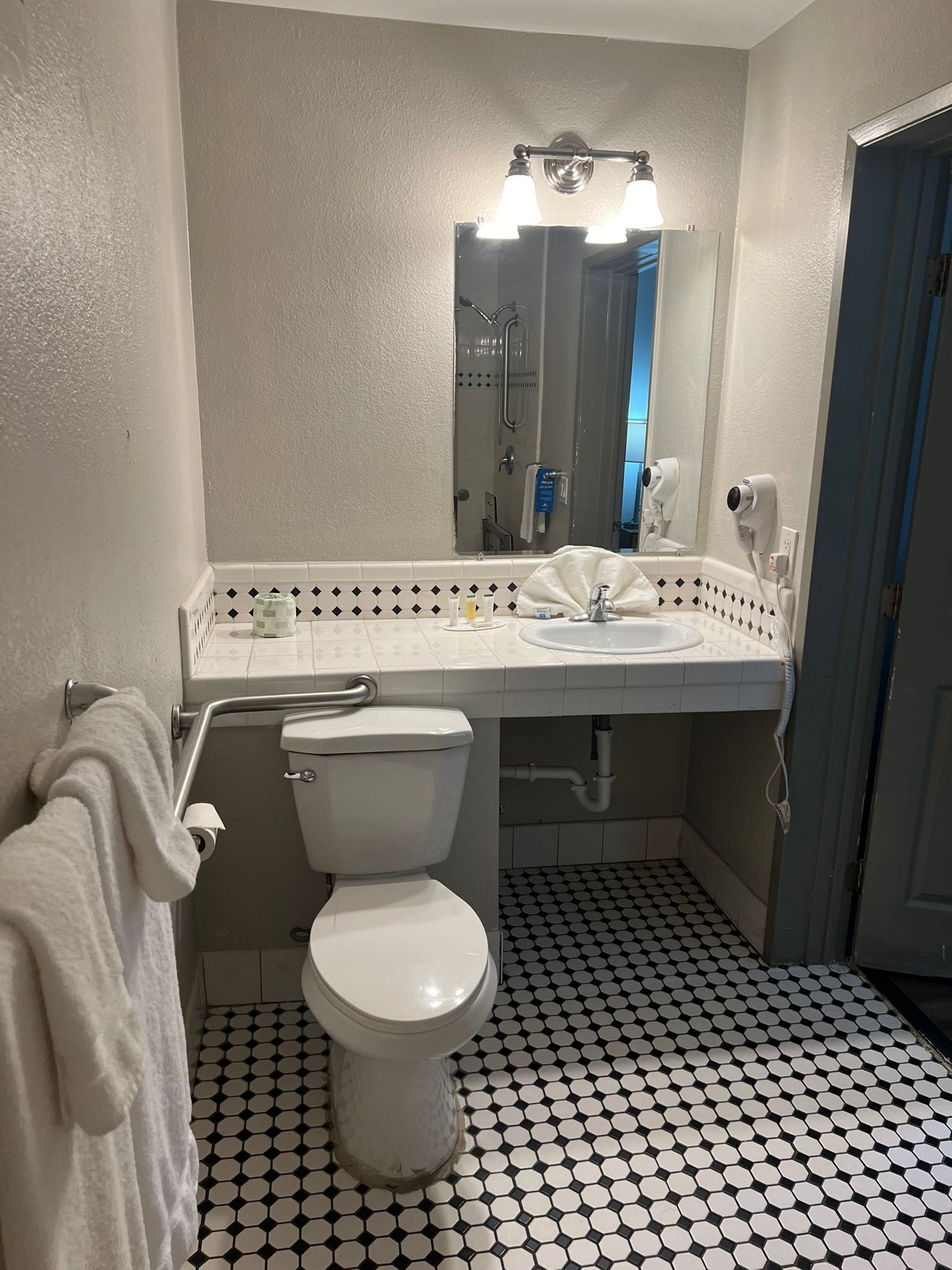 A bathroom with a toilet, sink, and mirror. There is a towel rack with a towel hanging on it. The floor has a black and white tile pattern.
