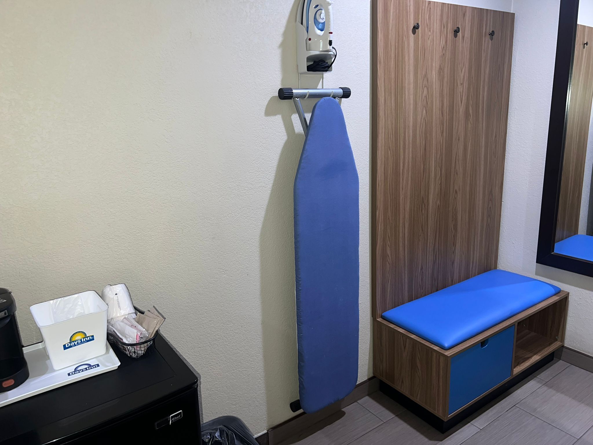Ein Hotelzimmer mit Holzschrank, blauer Sitzbank, Bügelbrett, Kaffeemaschine und Mülleimer.