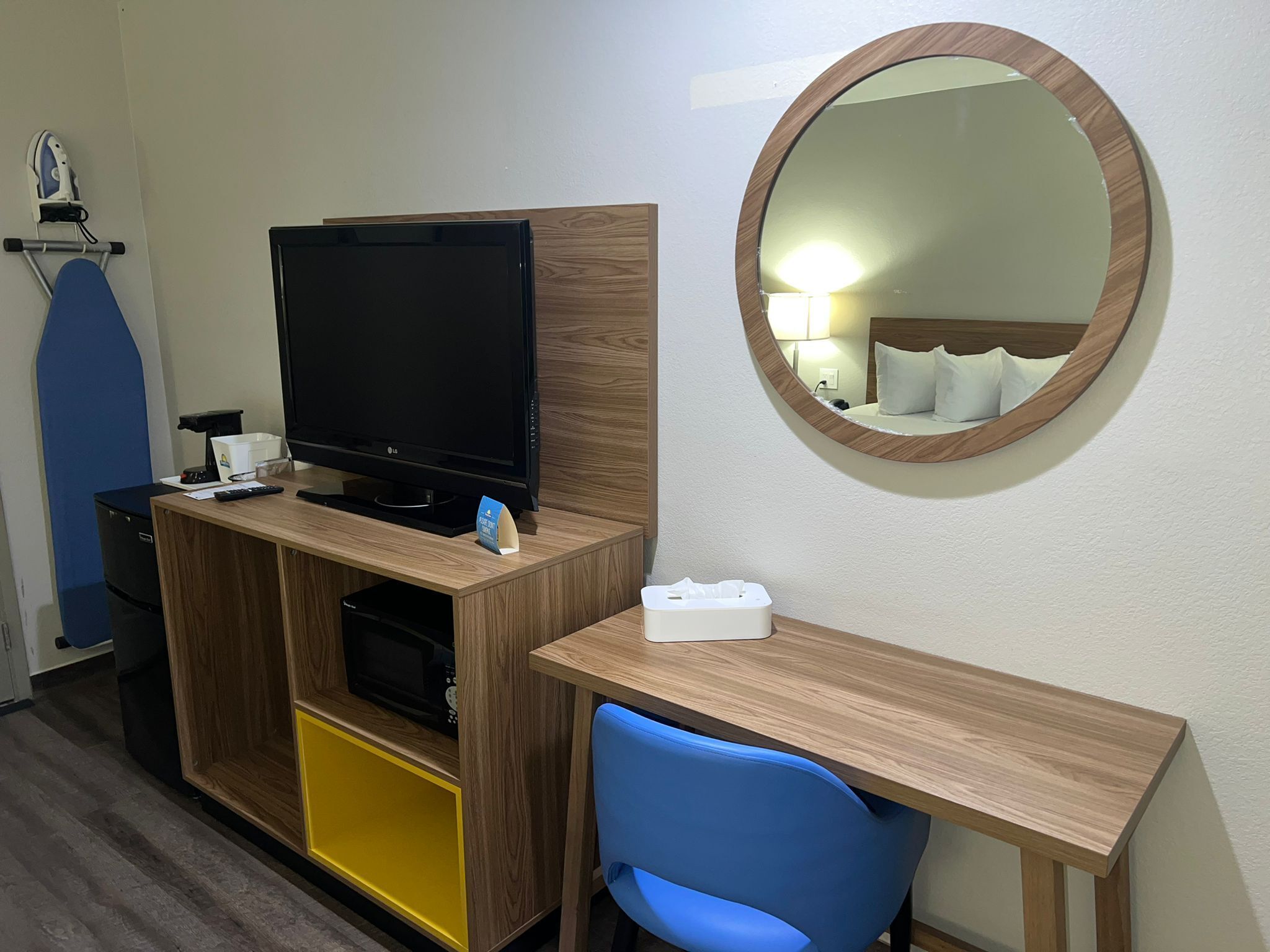 Habitación de hotel con escritorio de madera, televisión, microondas, una silla azul y un espejo redondo en la pared.