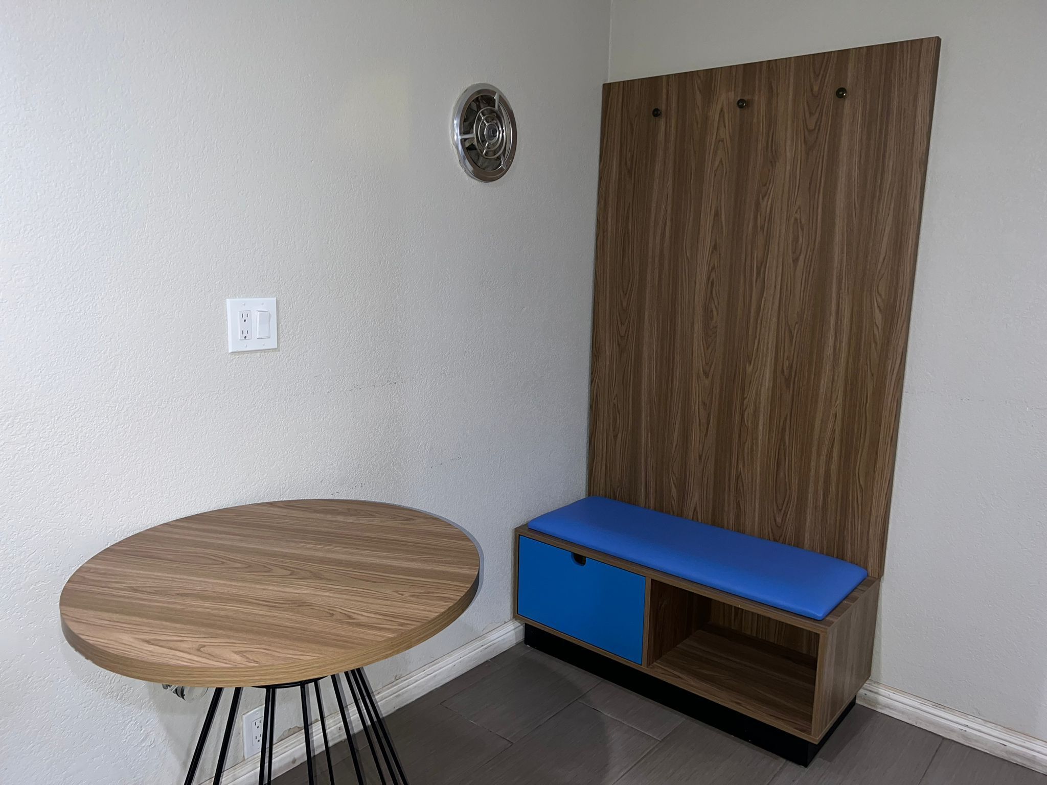 A corner room with a round wooden table, a blue bench with storage, a wooden wardrobe, and a wall socket.