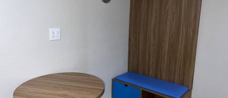 A corner room with a round wooden table, a blue bench with storage, a wooden wardrobe, and a wall socket.
