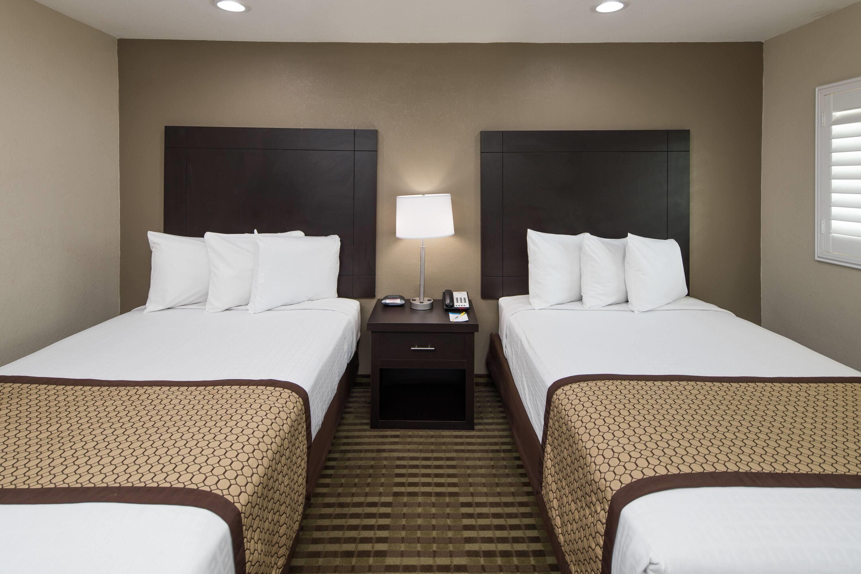A hotel room with two beds, a nightstand with a lamp, and a window with blinds.