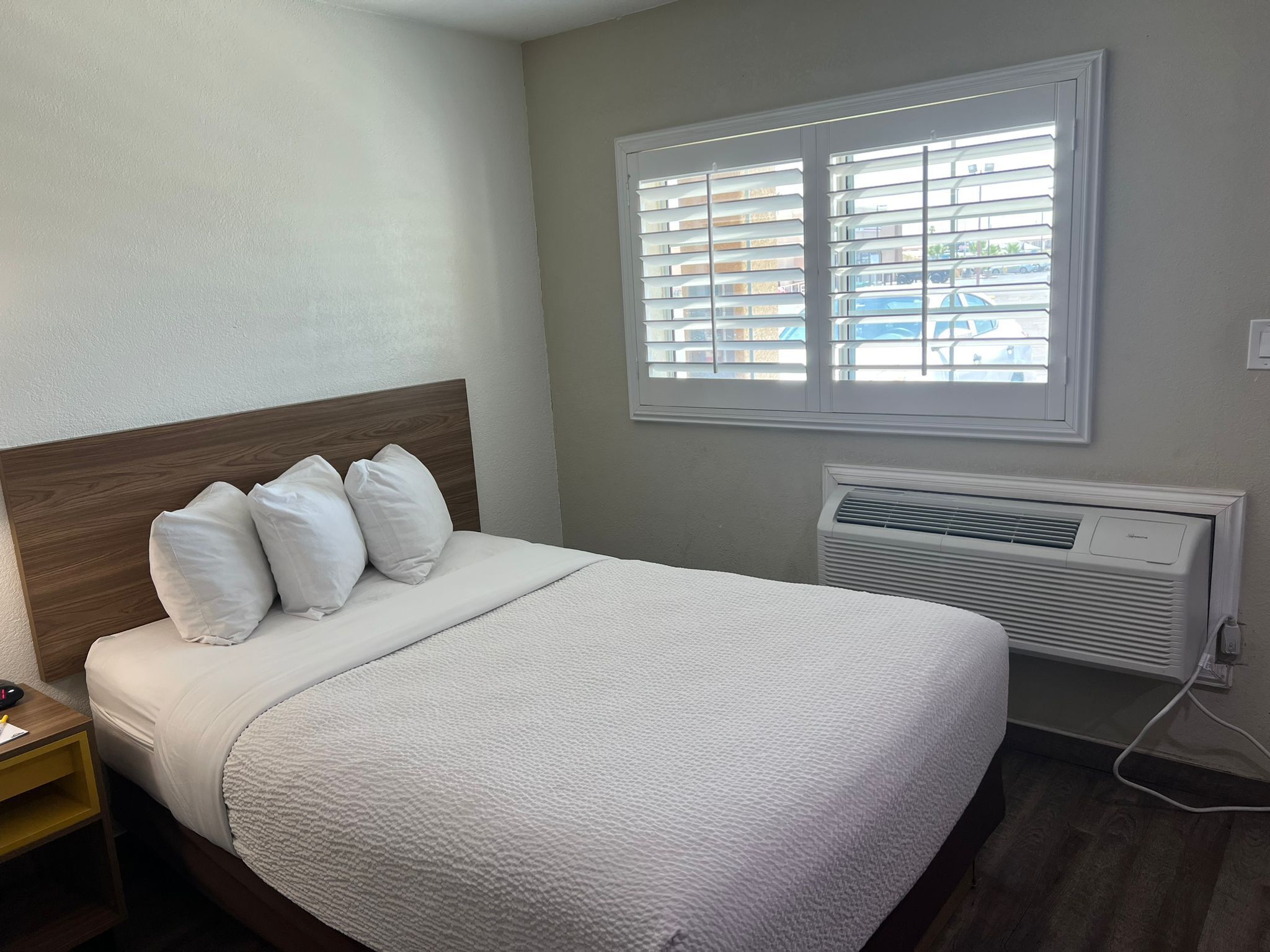 A neatly made bed with white bedding and pillows, a wooden headboard, and a window with blinds.