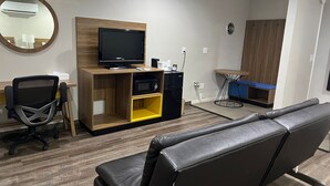 A modern hotel room with a wooden desk, a black office chair, a flat-screen TV, a microwave, a refrigerator, a small table with a blue bench, and a large mirror. - Days Inn by Wyndham El Centro (El Centro)