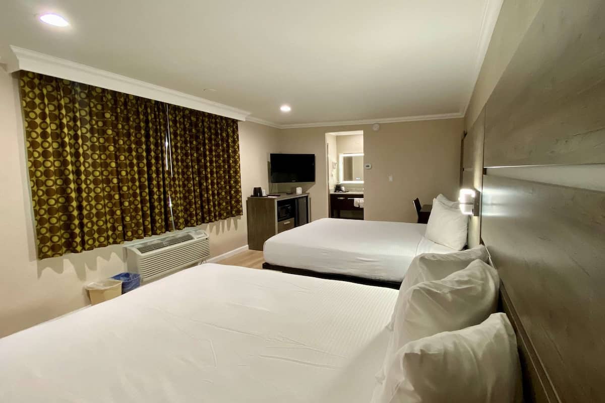 Habitación de hotel con dos camas, televisor, escritorio y baño visible a través de una puerta abierta.