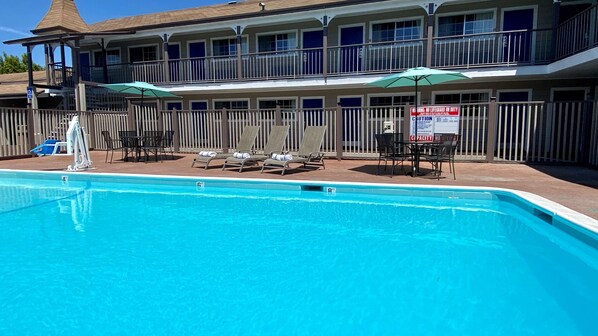 A pool area with lounge chairs and umbrellas, a lifeguard tower, and a building with multiple balconies.