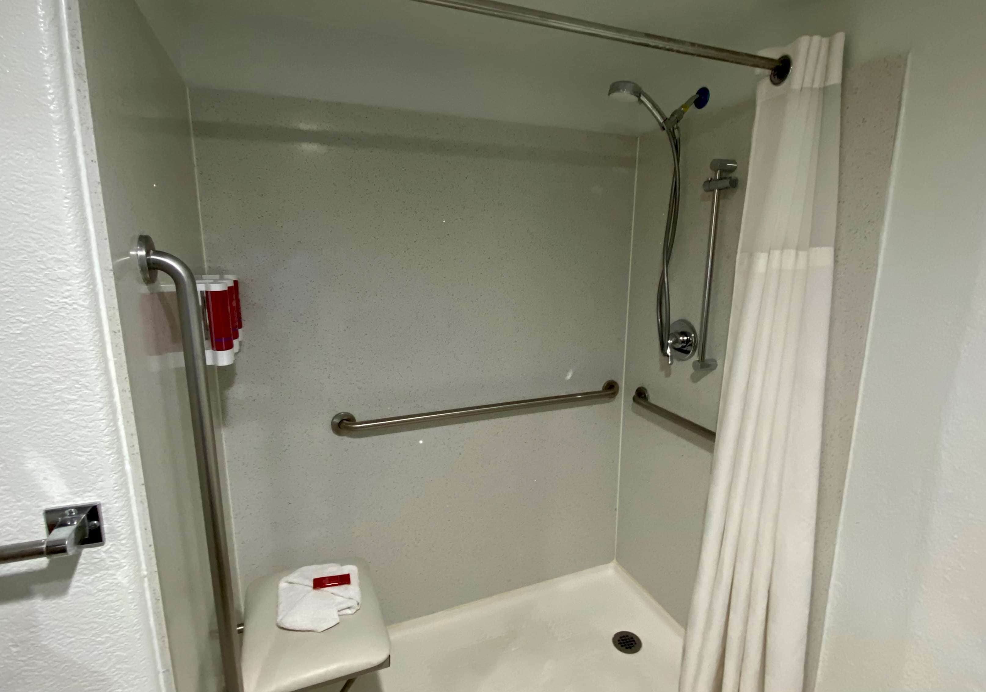 A small, enclosed shower area with a white shower curtain, a handheld showerhead, a grab bar, and a toilet with a white seat cover.