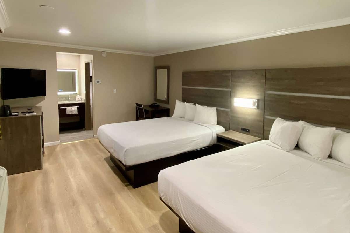 Habitación de hotel con dos camas, televisión, escritorio y baño visible a través de una puerta abierta.