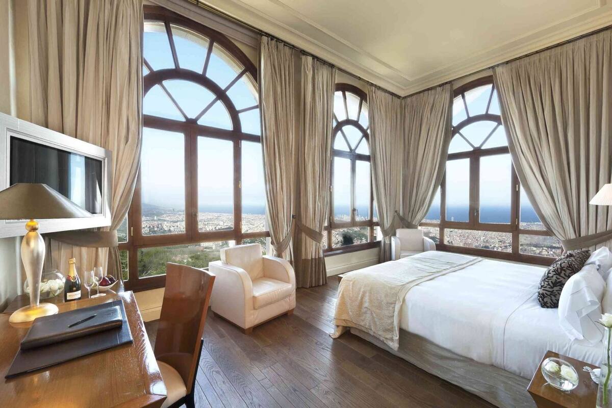 Quarto de hotel com uma cama grande, uma escrivaninha com luminária, uma cadeira e vista para a cidade através de amplas janelas.