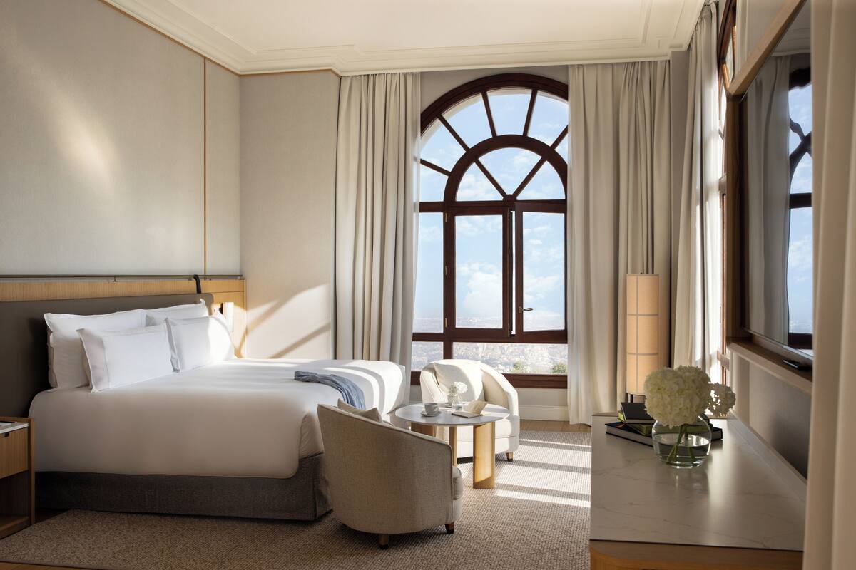 Quarto de hotel com uma cama grande, um sofá, uma mesa pequena e uma janela com cortinas.
