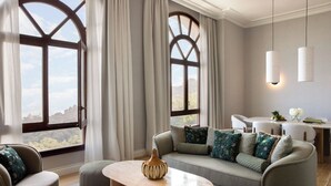 A modern living room with a sofa, ottoman, and coffee table. There are two windows with curtains, a dining area with a table and chairs, and pendant lights. - METT Barcelona GL – A Leading Hotel of the World (Barcelona)