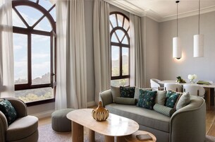 A modern living room with a sofa, ottoman, and coffee table. There are two windows with curtains, a dining area with a table and chairs, and pendant lights.
