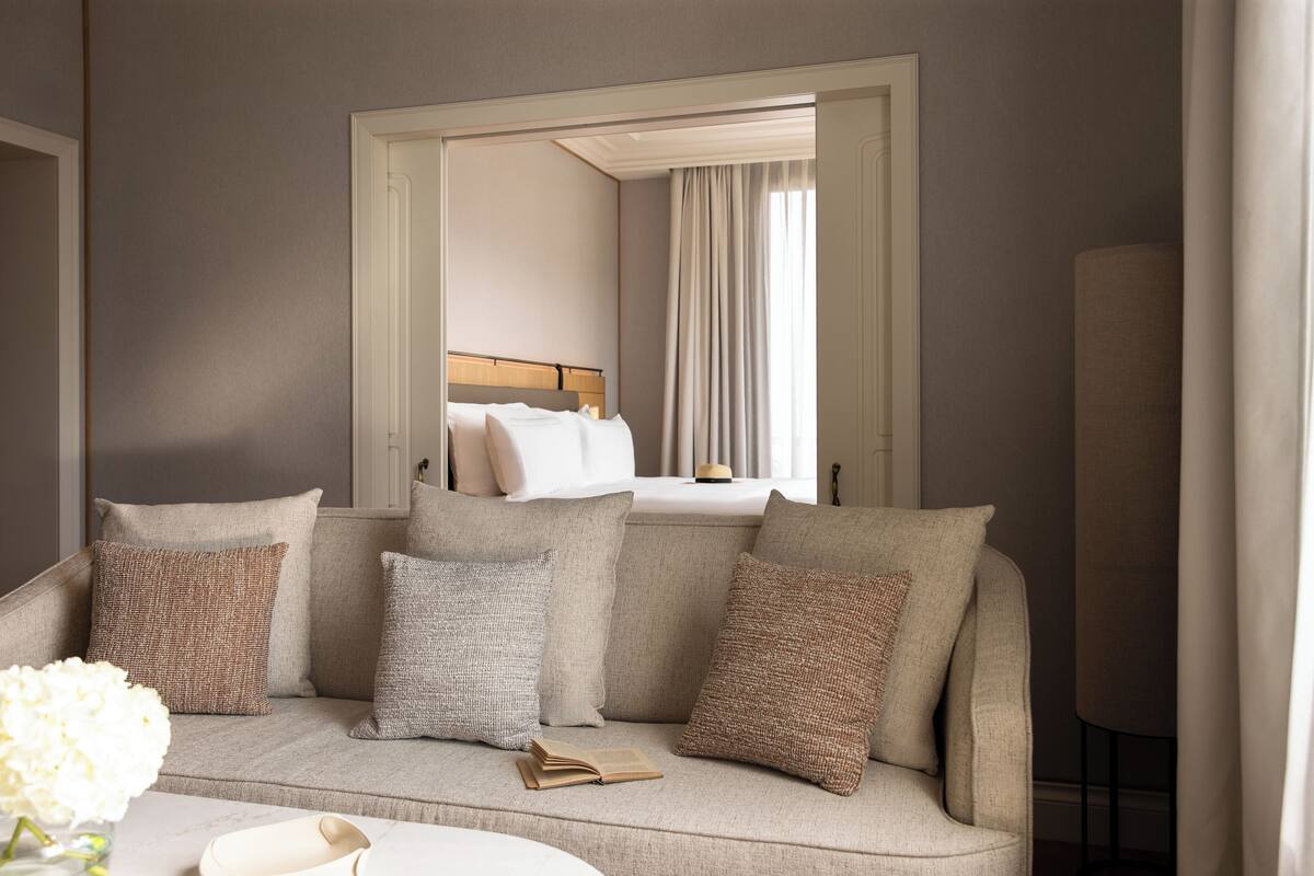 Quarto em tons neutros com um sofá, um espelho refletindo uma cama e um vaso com flores.